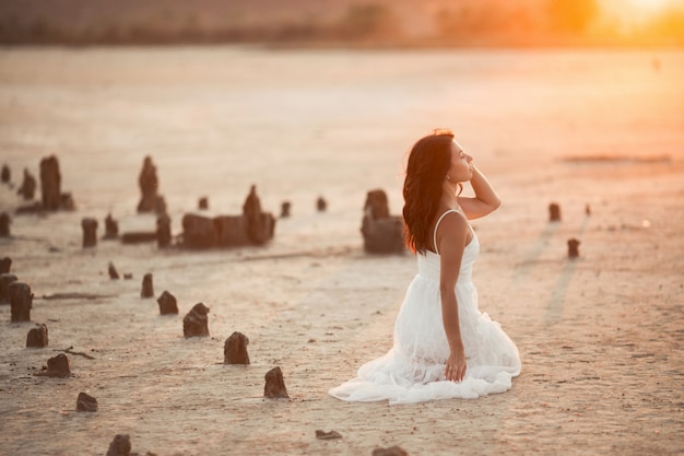 Vista lateral da menina morena que está sentado sobre os joelhos na areia por do sol
