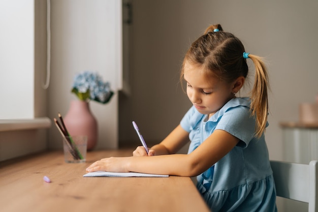 Vista lateral da menina primária focada aprendendo a escrever fazendo lição de casa sentada na mesa de casa pela janela. retrato de criança pré-escolar inteligente estudando sozinho fazendo anotações com caneta no quarto.