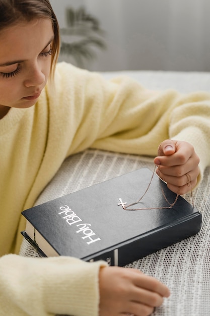 Vista lateral da menina rezando com a bíblia e o colar da cruz