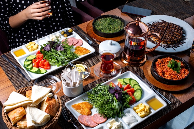 Vista lateral da mesa do café da manhã servida com vários alimentos ovos fritos salsichas queijo salada fresca sobremesa e chá