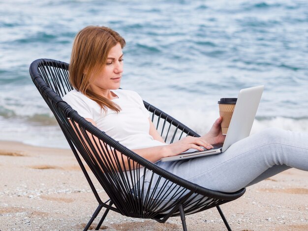 Vista lateral da mulher com café trabalhando na cadeira de praia no laptop