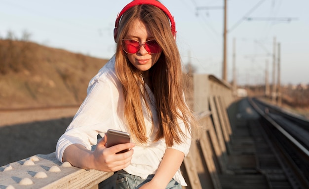 Vista lateral da mulher com óculos escuros e fones de ouvido segurando o smartphone