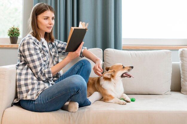 Vista lateral da mulher lendo livro no sofá com cachorro