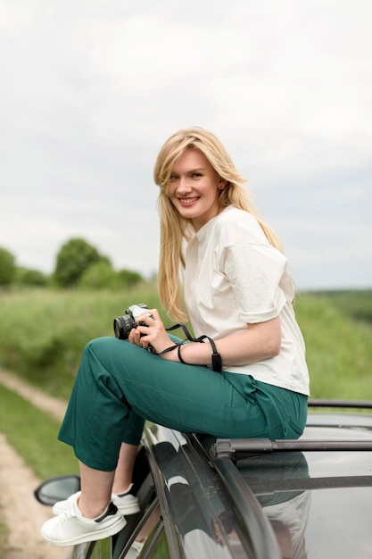 Foto grátis vista lateral da mulher sorridente posando em cima do carro enquanto segura a câmera