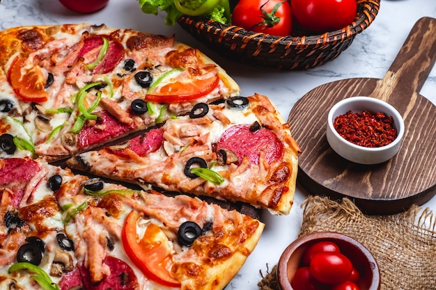 Vista lateral da pizza com salame presunto pimentão verde tomate azeitonas pretas e queijo em cima da mesa
