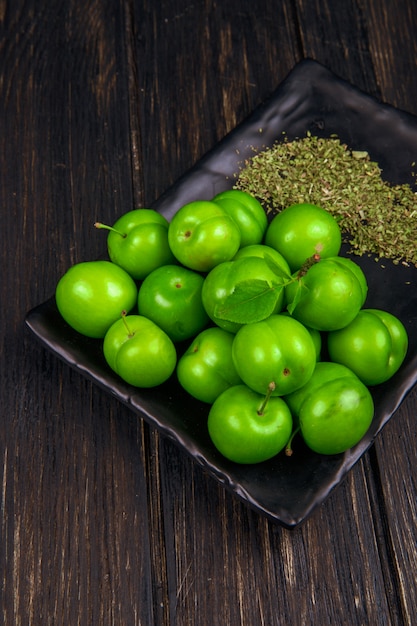 Vista lateral de ameixas verdes azedas com hortelã-pimenta seca em uma bandeja preta na mesa de madeira escura