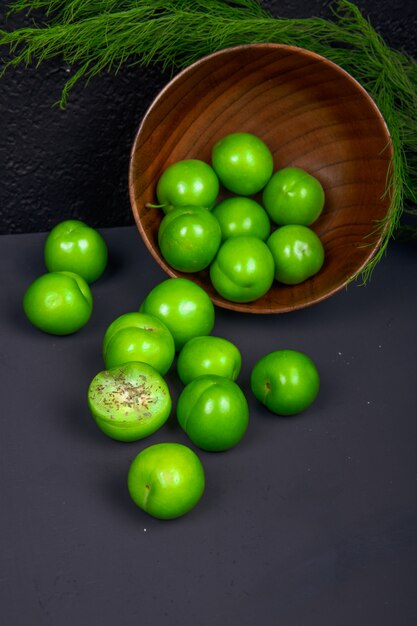 Vista lateral de ameixas verdes azedas espalhadas de uma tigela de madeira na mesa preta
