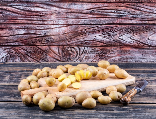 Vista lateral de batatas e fatias na tábua com descascador de batatas na mesa de madeira