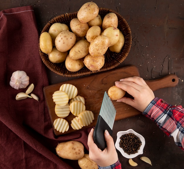 Vista lateral de batatas em uma cesta uma mulher corta batatas em uma placa com pimenta preta