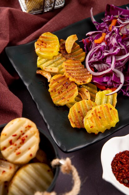 Vista lateral de batatas grelhadas com cebola roxa, repolho vermelho, pimentão laranja e flocos de pimenta seca em fundo cinza escuro