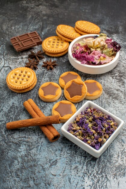 Vista lateral de biscoitos em uma bandeja de madeira e chocolates e tigelas de flores secas em um fundo cinza