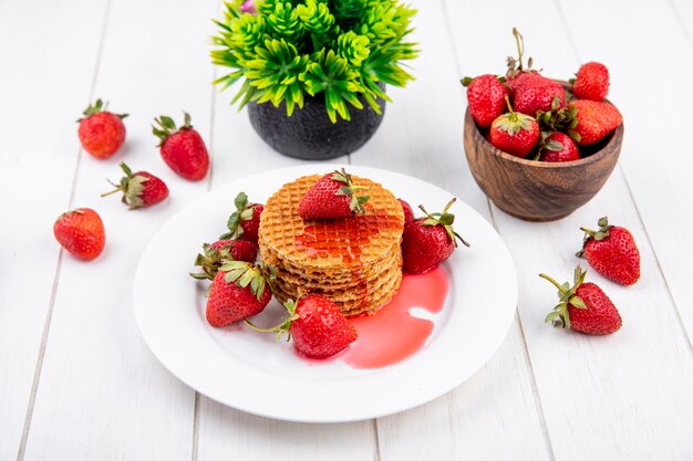 Vista lateral de biscoitos waffle com morangos no prato e na tigela com flores na madeira