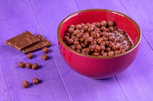 Foto grátis vista lateral de bolas de cereal de chocolate com leite em uma tigela e chocolate escuro no fundo de madeira roxo