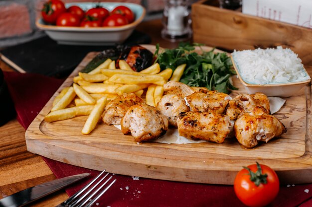 Vista lateral de carne de frango grelhado e legumes com batatas fritas e ervas em uma placa de madeira