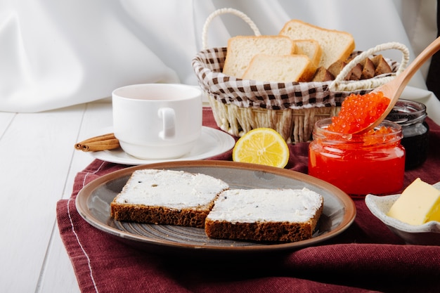 Vista lateral de caviar vermelho e preto com torradas em um prato com manteiga e uma xícara de chá numa toalha de mesa vermelha