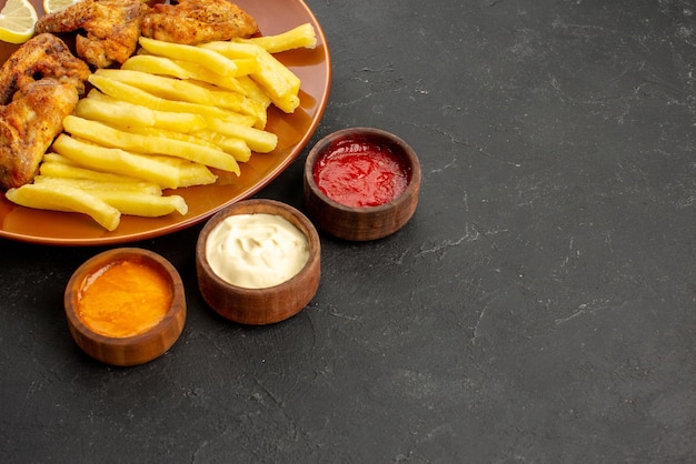 Vista lateral de close-up fastfood prato de laranja com asas de frango batatas fritas apetitosas e limão e três tigelas de diferentes tipos de molhos na mesa escura