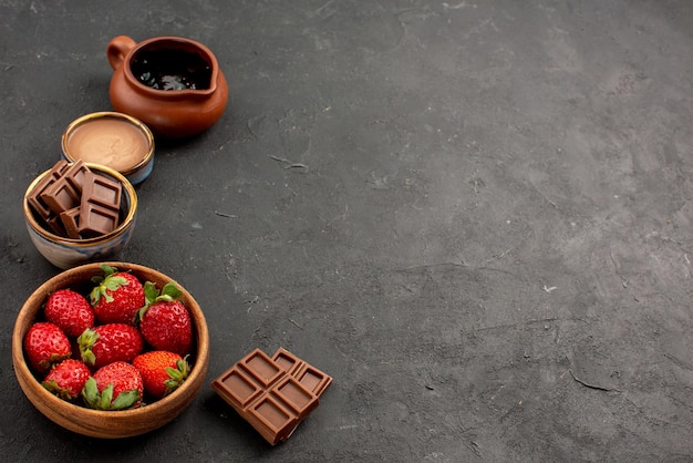 Vista lateral de close-up, morangos, morangos e creme de chocolate em tigelas e barras de chocolate no lado esquerdo da mesa escura