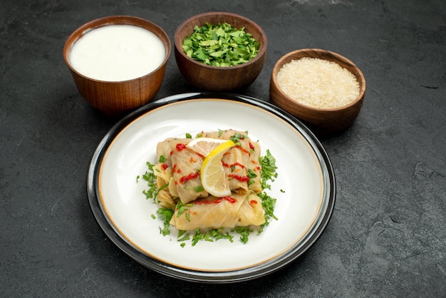 Vista lateral de close-up prato apetitoso repolho recheado com ervas de limão e molho no prato branco e tigelas com ervas de creme de leite e arroz no centro da mesa preta