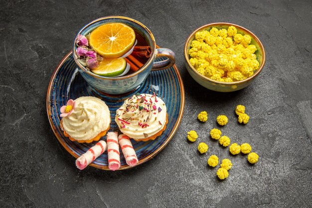 Vista lateral de close-up uma xícara de chá uma tigela de doces amarelos ao lado dos bolinhos apetitosos no prato e uma xícara de chá de ervas na mesa