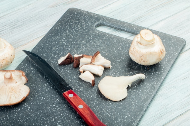 vista lateral de cogumelos frescos inteiros e fatiados com faca de cozinha em uma tábua preta na mesa rústica