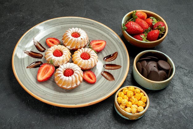 Vista lateral de cookies com biscoitos doces com chocolate e morango ao lado de tigelas de nozes de morango e chocolate em cima da mesa