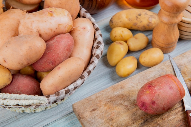 Vista lateral de diferentes tipos de batatas na cesta e corte um com faca na tábua com outros na mesa de madeira