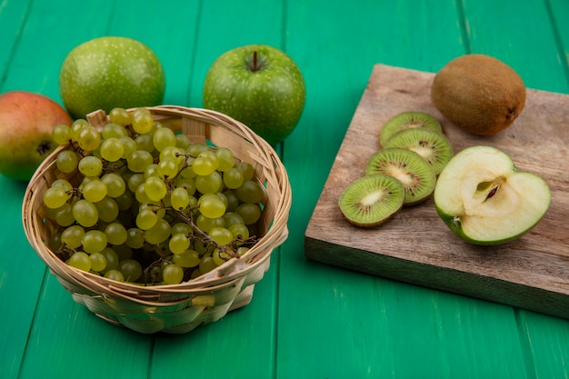 Vista lateral de fatias de kiwi em um carrinho com maçãs verdes e peras com uvas verdes em uma cesta sobre um fundo verde