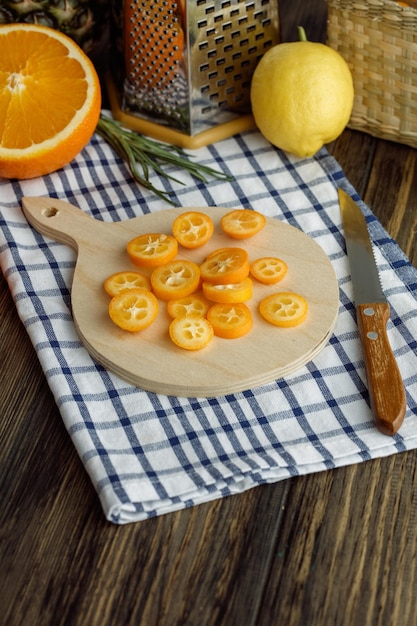 Foto grátis vista lateral de fatias de kumquat na tábua com faca raladora de cebolinha laranja cortada limão em pano xadrez em fundo de madeira