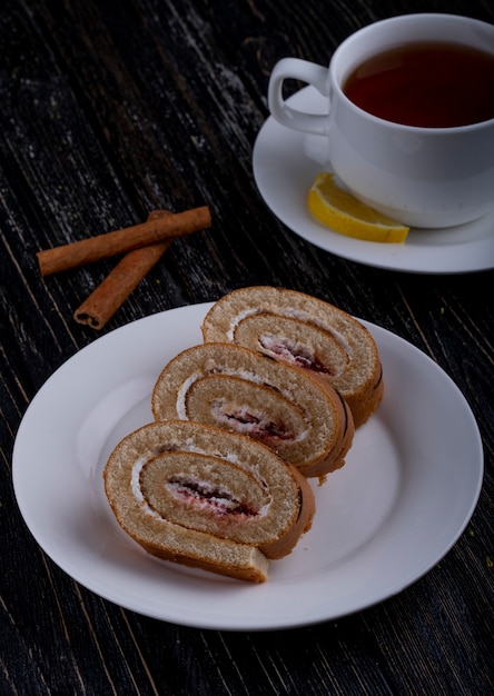 Vista lateral de fatias de rocambole com chantilly e geléia de framboesa em um prato servido com uma xícara de chá no rústico