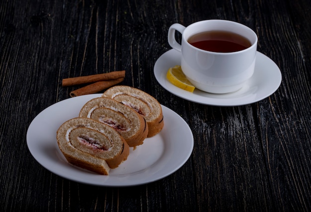 Vista lateral de fatias de rocambole com chantilly e geléia de framboesa em um prato servido com uma xícara de chá no rústico