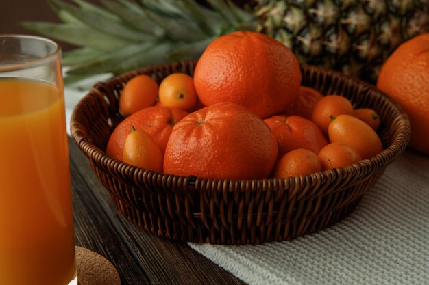 Vista lateral de frutas cítricas como tangerinas e kumquats na cesta de abacaxi laranja em pano com suco de laranja em fundo de madeira