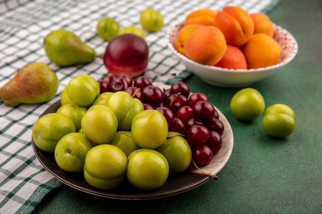 Vista lateral de frutas como ameixas cerejas damascos em prato e tigela com pêra e pêssego em pano xadrez sobre fundo verde
