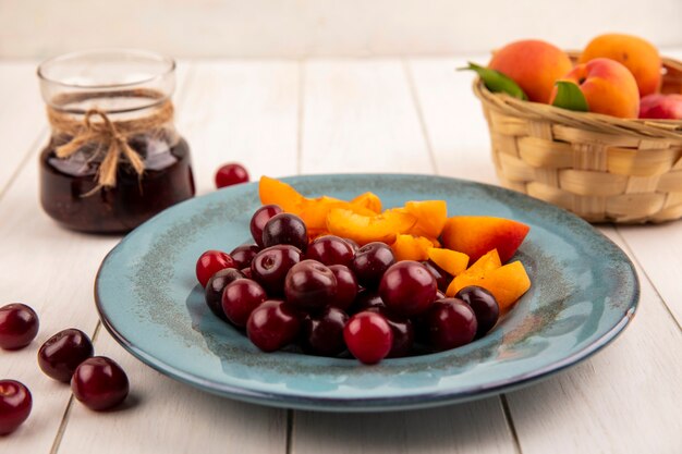 Vista lateral de frutas como cerejas e fatias de damasco no prato e cesta de damascos com geléia de morango no fundo de madeira