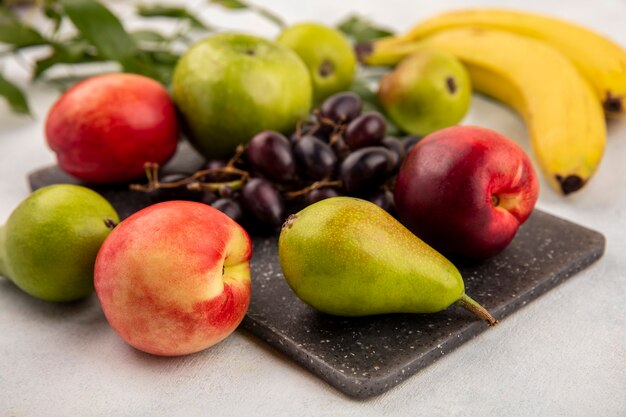 Vista lateral de frutas como pêra, maçã, uva, pêssego, numa tábua, com banana e folhas no fundo branco