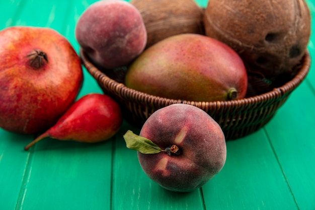 Vista lateral de frutas diferentes, como romã de manga e coco em um balde na superfície verde