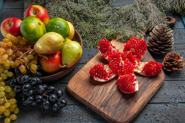 Vista lateral de frutas e cones uvas brancas e pretas maçãs limes peras em uma tigela de madeira ao lado de romã empilhada na mesa da cozinha e ramos de abeto com cones na mesa escura