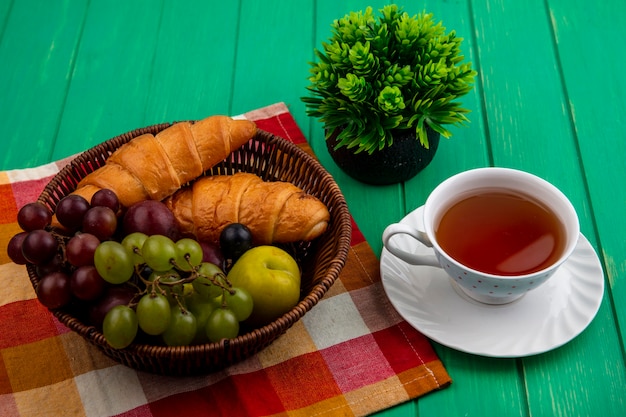 Vista lateral de frutas enquanto uva pluota bagas de abrunho com croissants em uma cesta em pano xadrez com uma xícara de chá e planta em fundo verde