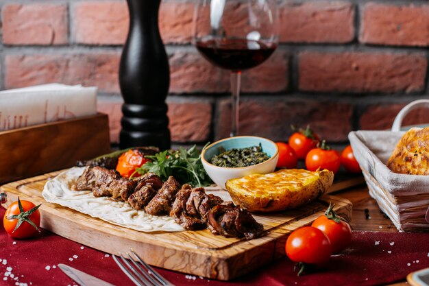 Vista lateral de kebab de cordeiro com batata cozida e legumes em uma placa de madeira