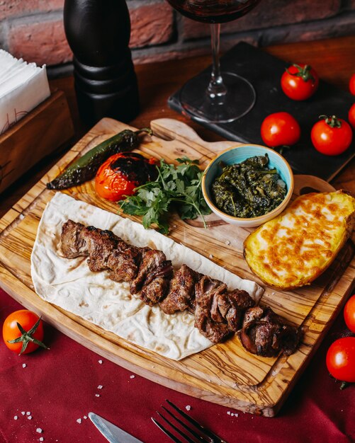 Vista lateral de kebab de cordeiro com batata cozida e legumes em uma placa de madeira
