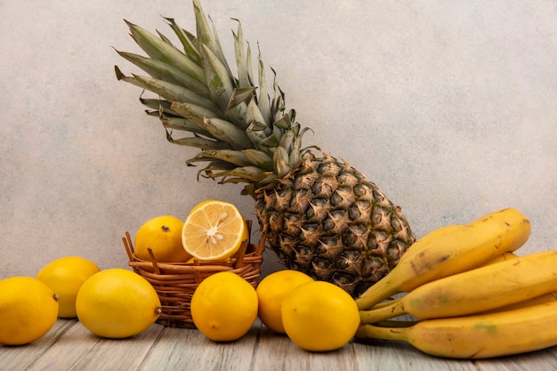 Vista lateral de limões amarelos em um balde com bananas e abacaxi, isolados em uma mesa de madeira cinza sobre uma superfície branca