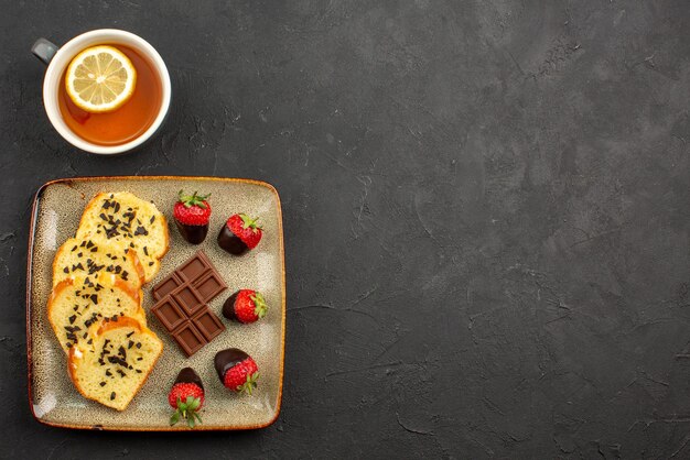 Vista lateral de longe xícara de chá com bolo cinza Prato de bolo com morangos cobertos de chocolate ao lado da xícara de chá com limão no lado esquerdo da mesa