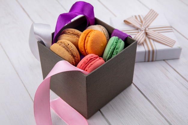 Vista lateral de macarons coloridos em uma caixa com arcos coloridos e papel de presente em uma superfície branca