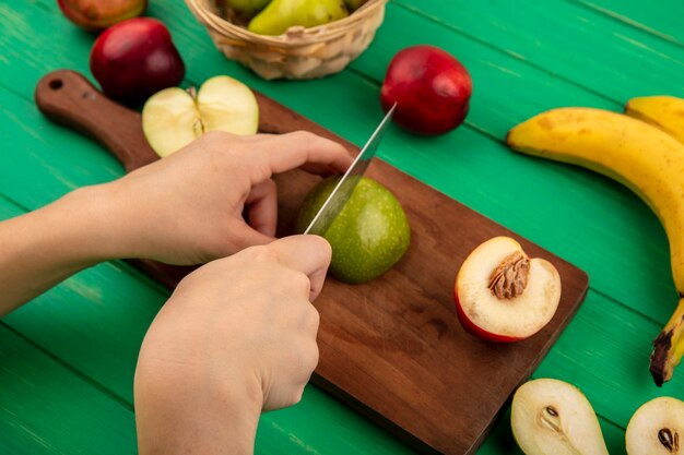 Vista lateral de mãos cortando maçã com faca e meio pêssego em uma tábua com banana e meio pêra cortada em fundo verde