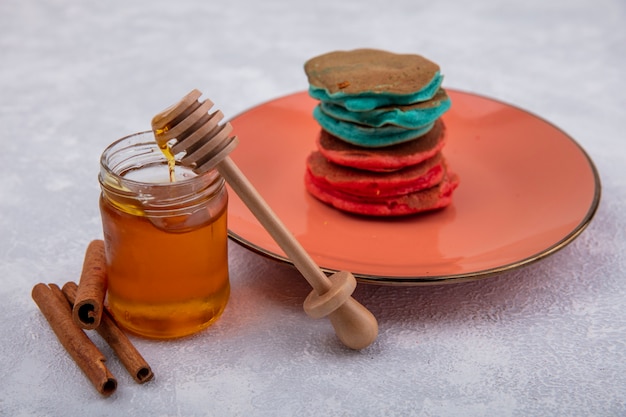Vista lateral de mel em um frasco com uma colher de pau de canela e panquecas coloridas em um prato de laranja em um fundo branco