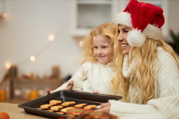 Foto grátis vista lateral de mulher loira vestindo chapéu de natal assando biscoitos com criança pequena olhando para a frente