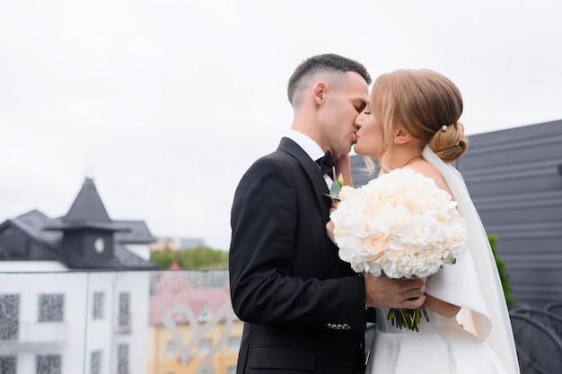 Foto grátis vista lateral de noivos fofos se beijando segurando buquês de flores em pé na varanda em tempo chuvoso no fundo da cidade antiga durante a primeira reunião emocional