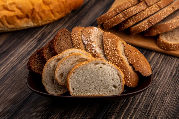 Vista lateral de pães como fatias de centeio-espiga marrom e outras brancas em uma tigela e na tábua de corte no fundo de madeira