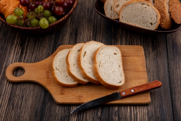 Vista lateral de pães em fatias de espiga marrom com sementes e brancas na tigela e na tábua com faca e cesta de uva para croissant no fundo de madeira