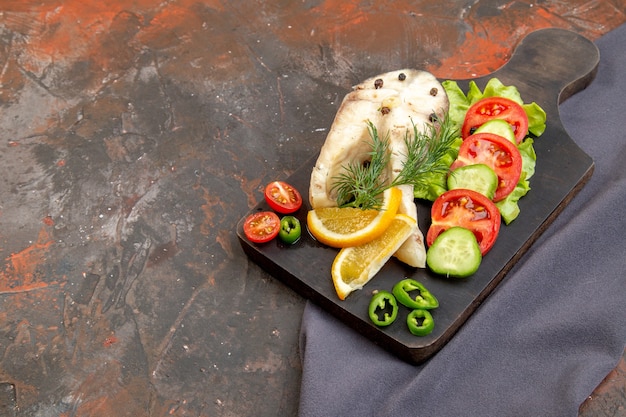 Vista lateral de peixes crus e alimentos frescos com pimenta em uma tábua de corte preta em uma toalha de cor escura em uma superfície de cor mista