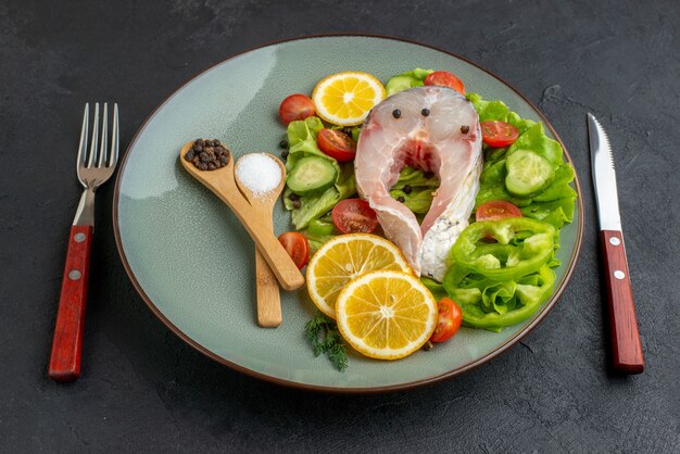 Vista lateral de peixes crus e vegetais frescos picados, fatias de limão, especiarias em um prato cinza e talheres colocados em uma superfície preta desgastada